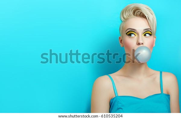 Fashionable girl\
with a stylish haircut inflates a chewing gum. The girl in the\
studio on a blue background. The girl\'s face with bright makeup and\
yellow with black shadows on the\
eyes.