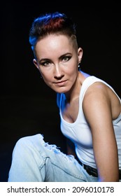 Fashionable Girl With Stylish Haircut Posing On Black Background With Trendy Colored Light. Face Girl With Bright Makeup In White T-shirt And Blue Jeans. Beautiful Confident Woman With Bold Eyeliner