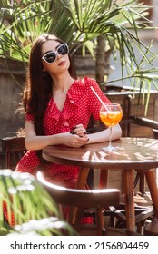 Fashionable Girl In Sunglasses, Straw Hat And Red Polka-dot Dress Sitting On The Summer Terrace With Aperol Spritz Drink. Young Nice Woman Portrait