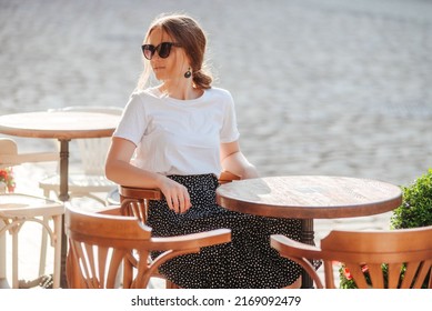 Fashionable girl wearing white t-shirt and polka-dot skirt sitting at table on the summer terrace of a cafe. Young woman portrait