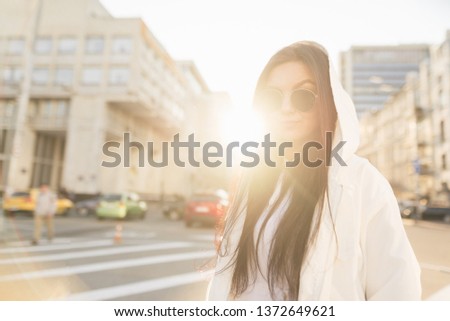 Fashionable girl in a white casual dress against the background of the sunset, street landscape. Stylish woman in sunglasses portrait with backlight on the street of megapolis.