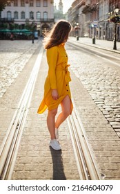 Fashionable Girl In Yellow Shirt Dress And White Sneakers Walk On Old City Street. Young European Woman Summer Portrait Outdoor