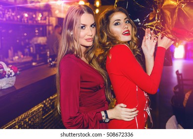 Fashionable girls, best friends posing in restaurant or  night club in stylish  sensual red dress with party balloons.  Drink cocktails, dancing. Bright make up. Wavy blonde hairstyle. 