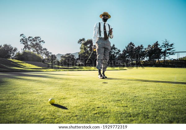 A fashionable good-looking mature bearded black\
guy in pants with suspenders and a straw hat, with a cigar and a\
golf club, is standing on the green grass of the lawn on a golf\
field on a warm evening