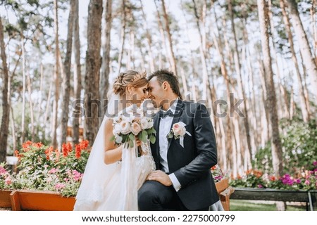 Fashionable groom and cute blonde bride in a white dress with a crown, a bouquet are hugging, laughing in the park, garden, forest outdoors. Wedding photography, portrait of smiling newlyweds.