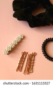 Fashionable hair accessories on pastel pink background; black scrunchie, goldenhair pins, coil hair tie and beret with pearls. Selective focus.