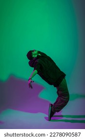 Fashionable handsome professional hip hop dancer man with a hat in a vest, jeans and sneakers dances in motion in a creative studio with green and purple light - Shutterstock ID 2278090587