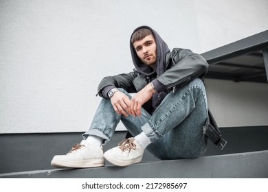 Fashionable handsome young hipster man wearing trendy clothes with jacket, hoodie, jeans and sneakers sitting near the white wall on the street - Powered by Shutterstock