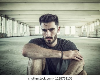 Fashionable Handsome Young Man Posing, Sitting on the Ground Inside an Empty Building While Looking at the Camera - Powered by Shutterstock