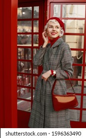 Fashionable Happy Smiling Blonde Woman Wearing Red Beret, Turtleneck, Checkered Coat, White Wrist Watch, Holding Stylish Leather Handbag,  Posing In Red Retro Street Phone Booth. 