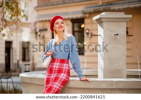 Fashionable happy smiling blonde woman wearing stylish red beret, blue cashmere sweater, checkered skirt, with white bag posing in street of European city. Copy, empty space for text