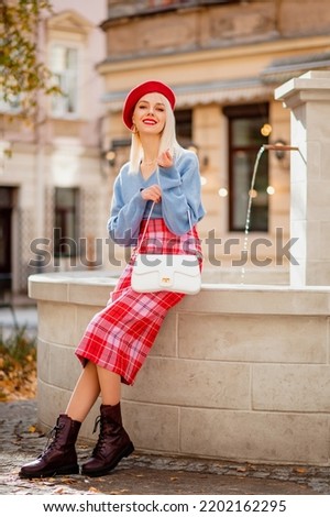 Fashionable happy smiling blonde woman wearing stylish red beret, blue cashmere sweater, checkered skirt, ankle boots, holding trendy white bag, posing in street of European city. Full-length portrait