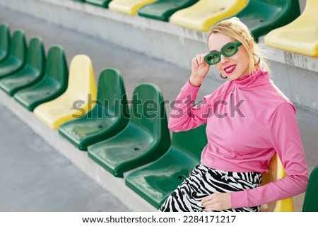 Fashionable happy smiling blonde woman wearing trendy green sunglasses, pink turtleneck, zebra print trousers, posing outdoors, on stadium seat. Copy, empty space for text