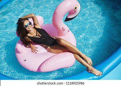 Fashionable, happy and smiling brunette model girl with perfect sexy body in stylish black bikini and glamorous sunglasses, posing on an inflatable pink flamingo at the swimming pool outdoors