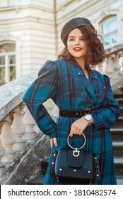 Fashionable happy smiling woman wearing stylish autumn outfit: faux leather beret, blue tartan dress, wrist watch, holding top handle black bag, posing in street