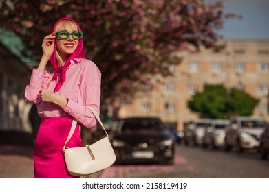Fashionable happy smiling woman wearing trendy green sunglasses, fuchsia color headscarf, classic pink shirt, satin mini skirt, holding white bag, walking in street of city. Copy, empty space for text