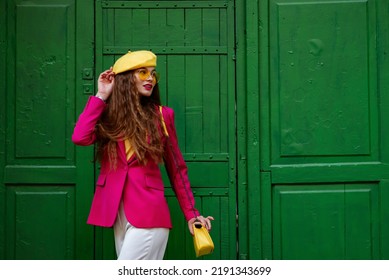 Fashionable happy smiling woman wearing trendy outfit with yellow sunglasses, beret, shoulder bag, pink fuchsia color blazer, posing near green door. Copy, empty space for text
