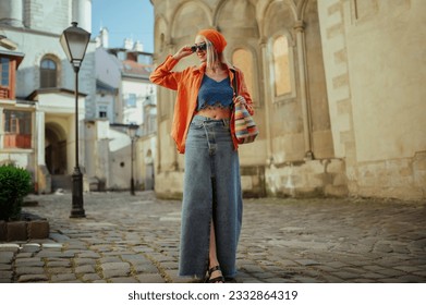 Fashionable happy smiling woman wearing trendy sunglasses, orange shirt, bandana, crop top, low waist denim maxi skirt, with wicker bag, posing in street of European city. Copy, empty space for text