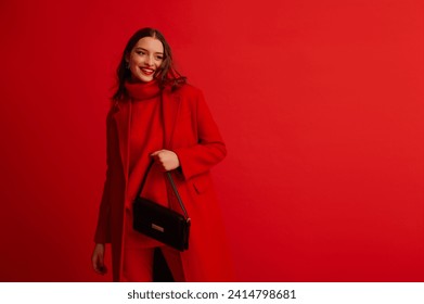 Fashionable happy smiling woman wearing trendy red midi coat, turtleneck sweater, holding black leather baguette bag, posing on red background. Studio fashion portrait. Copy, empty space for text - Powered by Shutterstock