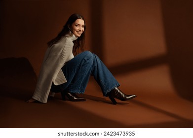 Fashionable happy smiling woman wearing oversized white sweater, wide leg jeans, leather cowboy boots, posing on brown background. Full-length studio fashion portrait. Copy, empty blank space for text - Powered by Shutterstock