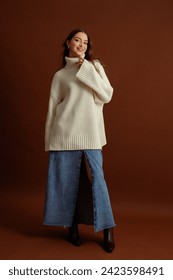 Fashionable happy smiling woman wearing oversized white turtleneck sweater, maxi blue denim skirt, pointed toe ankle leather boots, posing on brown background. Full-length studio fashion portrait