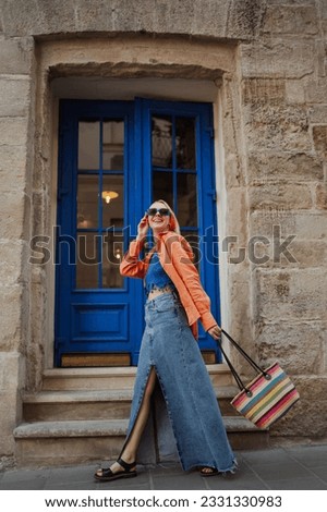 Fashionable happy smiling woman wearing trendy orange linen shirt, crochet crop top, maxi denim skirt, blue sunglasses, holding striped wicker tote bag, walking in street. Outdoor full-length portrait