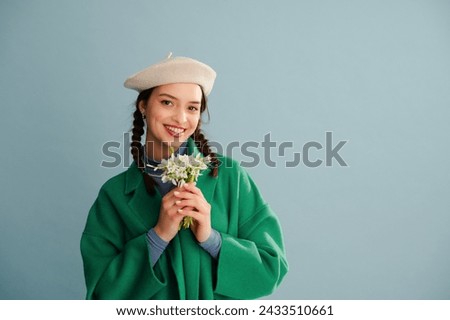 Fashionable happy smiling woman wearing white beret, trendy green coat, holding spring bouquet of snowdrops, posing on blue background. Studio portrait. Copy, empty, blank space for text