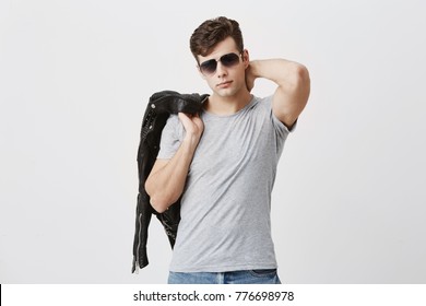 Fashionable hipster with dark hair posing in stylish eyewear, casually dressed, thrown black leather jacket over his shoulder. Muscular fit male model looking with appeal and confidence at camera. - Powered by Shutterstock