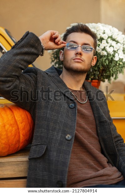 Fashionable hipster man with coat and sunglasses\
sits near a vintage yellow car with flowers and pumpkin. Autumn\
men’s style clothes