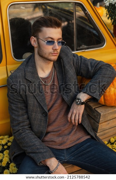 Fashionable\
hipster man with hairstyle and trendy vintage sunglasses in fashion\
coat, jeans and sweatshirt with luxury watch sits near yellow car\
countryside. Men’s autumn style look\
clothes
