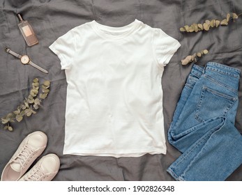 Fashionable look with white empty t-shirt, jeans and white sneakers. Top view of white blank t-shirt with short sleeves over gray bed linen. Mock up for t-shirt print design.