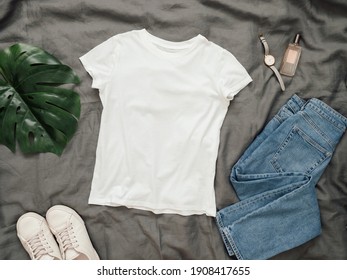Fashionable look with white empty t-shirt, jeans and white sneakers. Top view of white blank t-shirt with short sleeves over gray bed linen. Mock up for t-shirt print design.