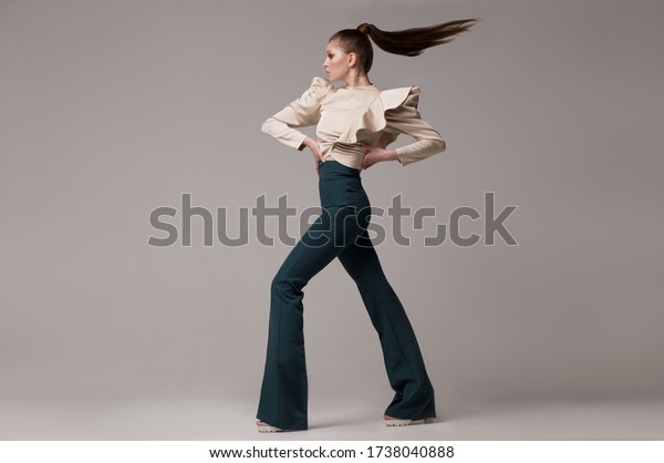 Fashionable look, a woman in long green flared\
trousers, a silk beige blouse with wide magnificent sleeves. A\
model poses in a studio on a light background, her hair is flying.\
Fashion clothes.