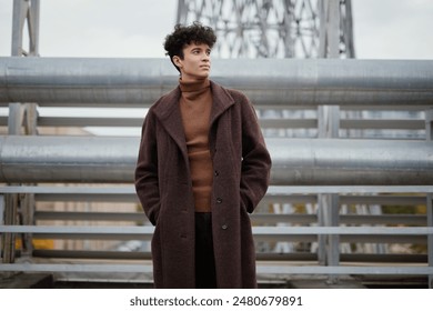 Fashionable man in brown coat and turtleneck standing in front of steel bridge with railing - Powered by Shutterstock