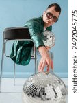 Fashionable man with cerebral palsy wearing trendy green color sunglasses, sequin jacket, jeans, sneakers, holding disco ball, sitting on chair. Full-length studio portrait