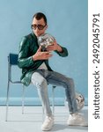 Fashionable man with cerebral palsy wearing trendy green color sunglasses, sequin jacket, jeans, sneakers, holding disco ball, sitting on chair. Full-length studio portrait