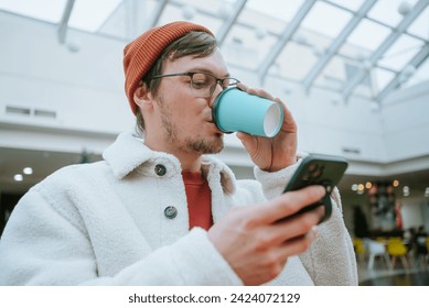 A fashionable man in a winter coat and knit beanie enjoys a coffee and checks his phone in a modern shopping center atrium.  - Powered by Shutterstock