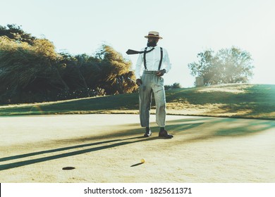 A Fashionable Mature Bearded Black Guy In A Hat, White Shirt, And Trousers With Suspenders Is Holding In His Hands A Cigar And A Golf Club While Standing On A Golf Field Next To The Ball And The Hole