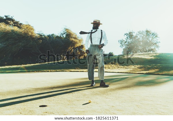 A fashionable mature bearded black guy in a hat,\
white shirt, and trousers with suspenders is holding in his hands a\
cigar and a golf club while standing on a golf field next to the\
ball and the hole