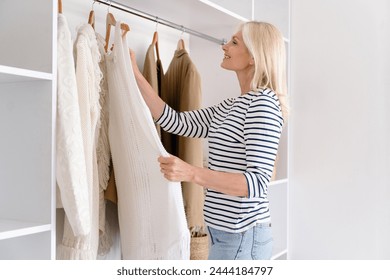 Fashionable mature woman standing with dress in small boutique, choosing new stylish outfit. Mid age female looking perfect casual clothes in wardrobe, hold hanger with apparel in home dressing room - Powered by Shutterstock