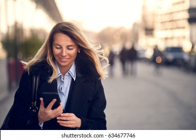 Fashionable Middle-aged Woman In Black Coat And Blond Hair Busy With Her Mobile Phone While Walking A City Street