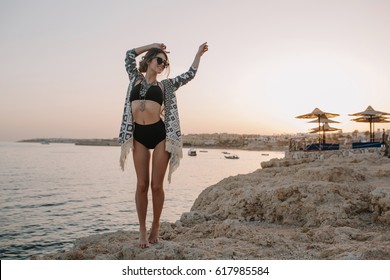 Fashionable Model Posing On Beach, On Sunset, Girl On Vacation. Sexy Woman Wearing Black Bikini, Swimsuit With Hight Waist, Cardigan, Cape With Ornaments.