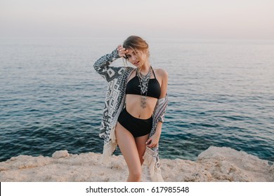 Fashionable Model Posing On Rock Beach, On Sunset, Near Sea, Ocean. Sexy Woman Wearing Black Bikini, Swimsuit With Hight Waist, Cardigan, Cape With Ornaments.