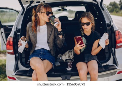 Fashionable Mother With Daughter. Family Is Sitting In The Trunk. Girl In A Black T-shirt. Ladies Eating A Donuts