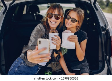 Fashionable Mother With Daughter. Family Is Sitting In The Trunk. Girl In A Black T-shirt. Ladies Eating A Donuts