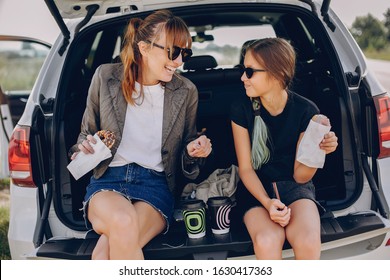 Fashionable Mother With Daughter. Family Is Sitting In The Trunk. Girl In A Black T-shirt. Ladies Eating A Donuts