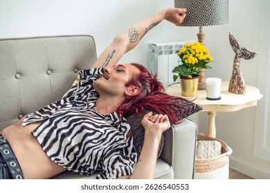 Fashionable non-binary person with pink hair and tattoos relaxing on a comfortable sofa at home, enjoying their free time in a body positive and gender-fluid way