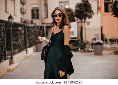 Fashionable pale brunette in long green dress, black jacket and sunglasses, standing on street during daytime against background of light city building