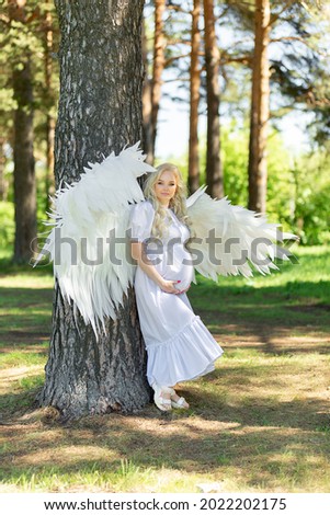 Fashionable portrait of a happy pregnant woman in an elegant white dress with angel wings in the city park