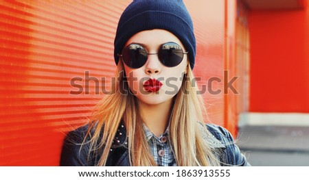 Fashionable portrait of stylish beautiful blonde young woman model blowing her lips with red lipstick sends sweet kiss in round sunglasses, black rock style leather jacket, hat on city street